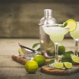 Dwa orzeźwiające drinki margarita z solą na brzegach kieliszków, ozdobione limonką i miętą. Obok shaker i świeże limonki. Idealny drink margarita przepis.