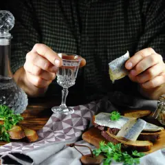 Śledź vs ogórek: jakie zakąski najlepiej pasują do czystej wódki? Na stole wódka, śledzie na chlebie, ogórki w słoiku i natka pietruszki.