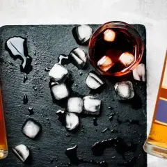 Dwie butelki whisky, szklanka z lodem i rozlaną wodą. Metaliczny posmak i pieczenie to objawy niskiej jakości whisky zbożowej.