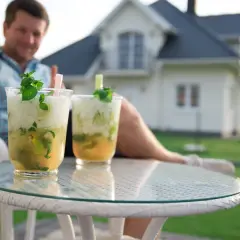 Jak przygotować drinki w dzbankach na domową imprezę? Dwa orzeźwiające mohito z miętą i limonką na stoliku, w tle mężczyzna i dom.