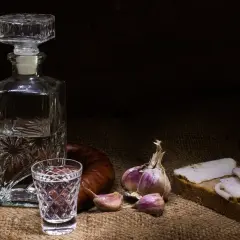 Wódka smakowa w karafce i kieliszku, obok kiełbasa, czosnek i chleb ze smalcem. Widoczne objawy utlenienia wódki smakowej po długim staniu w słońcu.