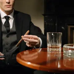 Mężczyzna w garniturze trzyma cygaro, obok szklanka whisky i karafka. Dobór whisky do cygar: intensywność i profil aromatu.