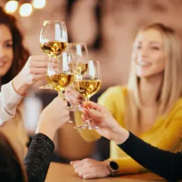 Przyjaciele wznoszą toasty kieliszkami białego wina, zastanawiając się, ile kosztuje butelka wina w restauracji.