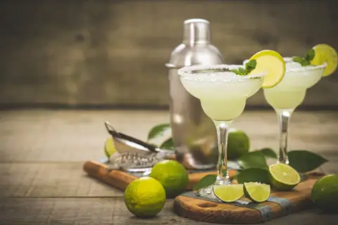 Dwa orzeźwiające drinki margarita z solą na brzegach kieliszków, ozdobione limonką i miętą. Obok shaker i świeże limonki. Idealny drink margarita przepis.