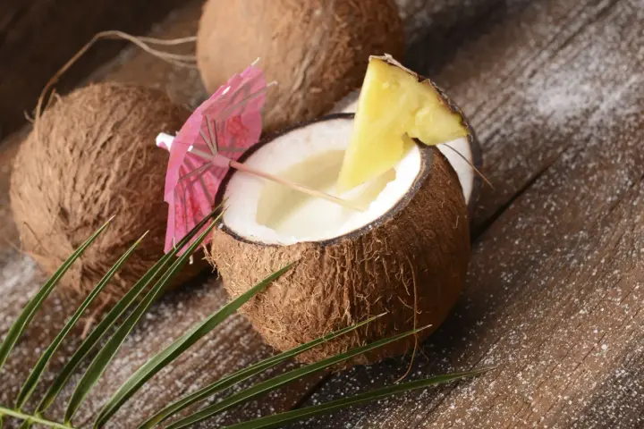 Orzeźwiający koktajl Malibu z mlekiem, podany w połówce kokosa, ozdobiony ananasem i parasolką. W tle inne kokosy i liście palmy.