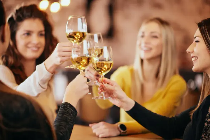 Przyjaciele wznoszą toasty kieliszkami białego wina, zastanawiając się, ile kosztuje butelka wina w restauracji.