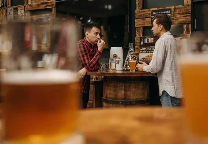 Dwóch mężczyzn rozmawia przy barze, na którym stoi kufel i szklanka piwa. Dyskutują, ile schodzi jedno piwo, podczas gdy w tle widać drewniane dekoracje i plakaty.