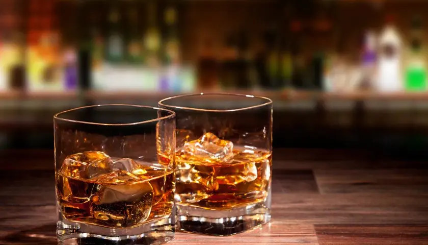 Dwa szklanki whisky z lodem na drewnianym blacie. W tle rozmyty bar, sugerujący atmosferę, która napędza przyczyny popularności whisky rzemieślniczej.