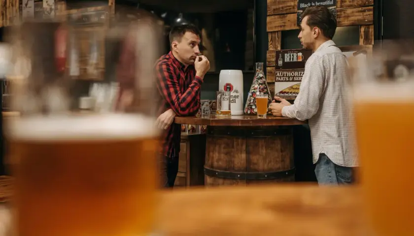 Dwóch mężczyzn rozmawia przy barze, na którym stoi kufel i szklanka piwa. Dyskutują, ile schodzi jedno piwo, podczas gdy w tle widać drewniane dekoracje i plakaty.
