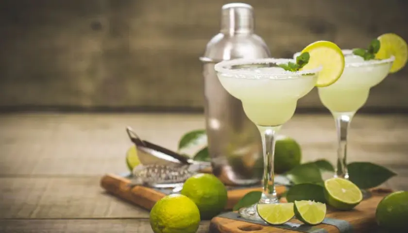 Dwa orzeźwiające drinki margarita z solą na brzegach kieliszków, ozdobione limonką i miętą. Obok shaker i świeże limonki. Idealny drink margarita przepis.