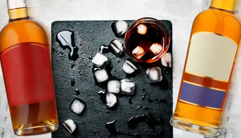 Dwie butelki whisky, szklanka z lodem i rozlaną wodą. Metaliczny posmak i pieczenie to objawy niskiej jakości whisky zbożowej.
