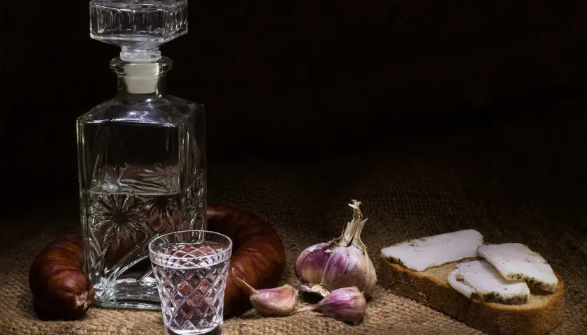 Wódka smakowa w karafce i kieliszku, obok kiełbasa, czosnek i chleb ze smalcem. Widoczne objawy utlenienia wódki smakowej po długim staniu w słońcu.