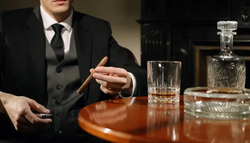 Mężczyzna w garniturze trzyma cygaro, obok szklanka whisky i karafka. Dobór whisky do cygar: intensywność i profil aromatu.