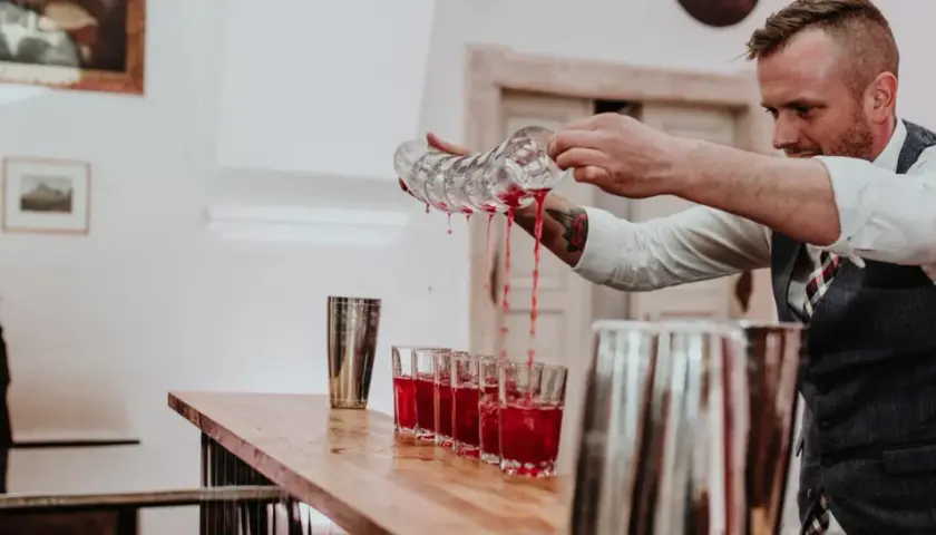 Barman nalewa czerwony napój do kieliszków na weselu, obok podekscytowana panna młoda.