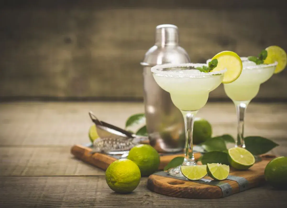 Dwa orzeźwiające drinki margarita z solą na brzegach kieliszków, ozdobione limonką i miętą. Obok shaker i świeże limonki. Idealny drink margarita przepis.