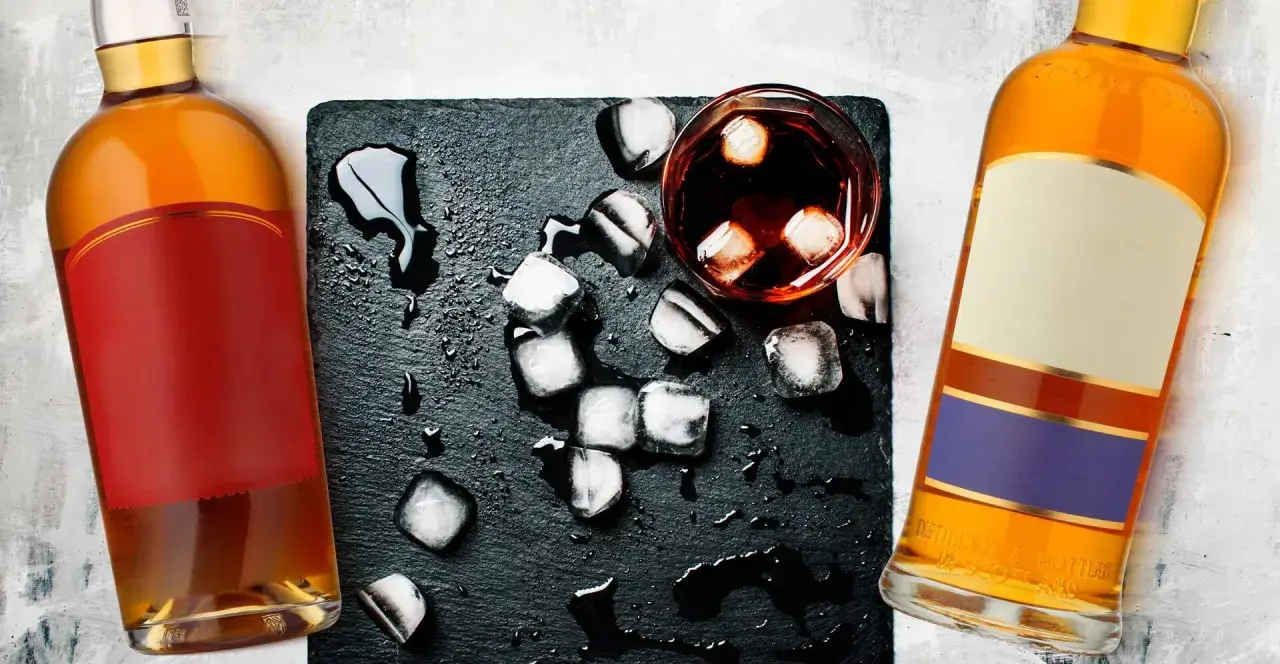 Dwie butelki whisky, szklanka z lodem i rozlaną wodą. Metaliczny posmak i pieczenie to objawy niskiej jakości whisky zbożowej.