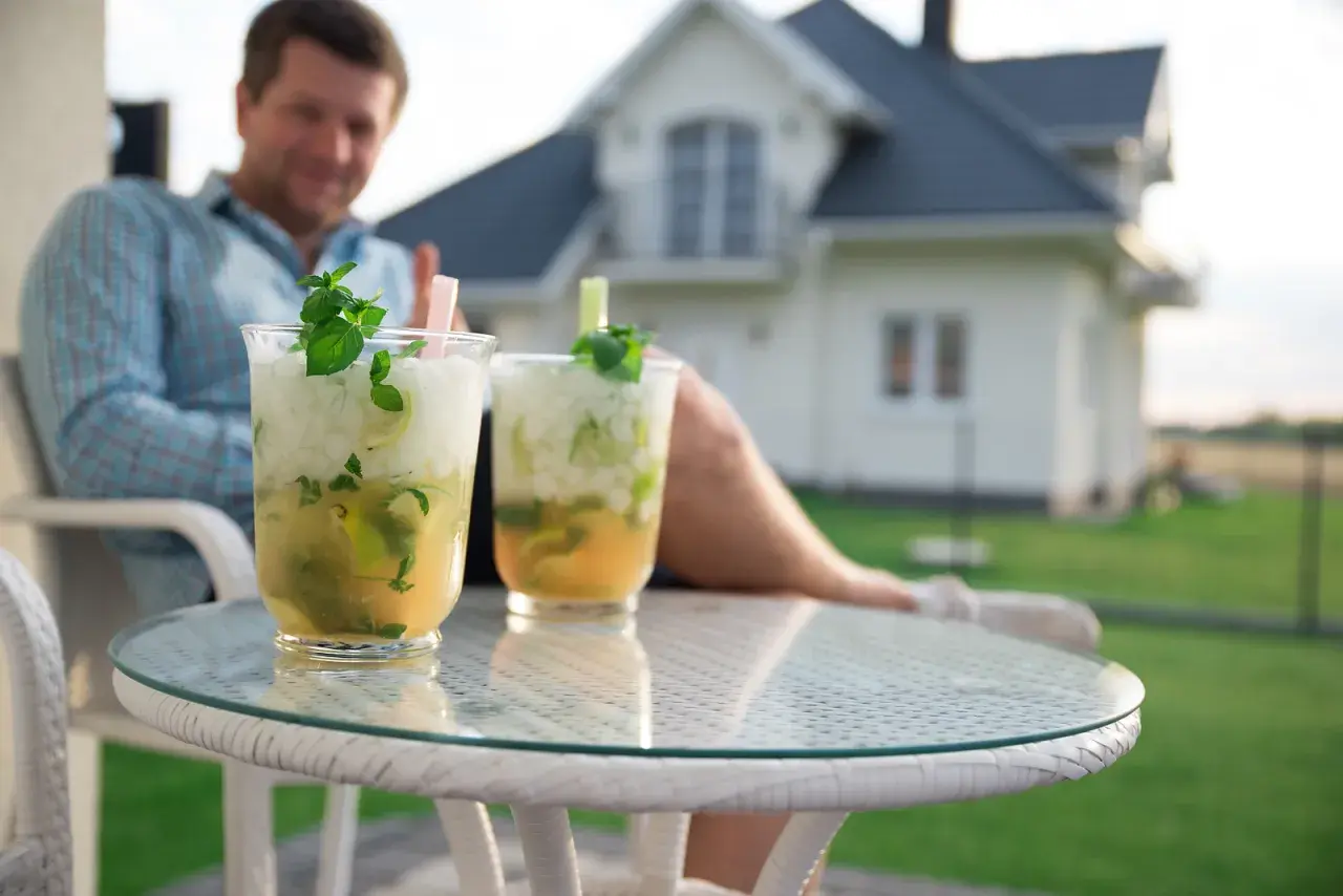 Jak przygotować drinki w dzbankach na domową imprezę? Dwa orzeźwiające mohito z miętą i limonką na stoliku, w tle mężczyzna i dom.