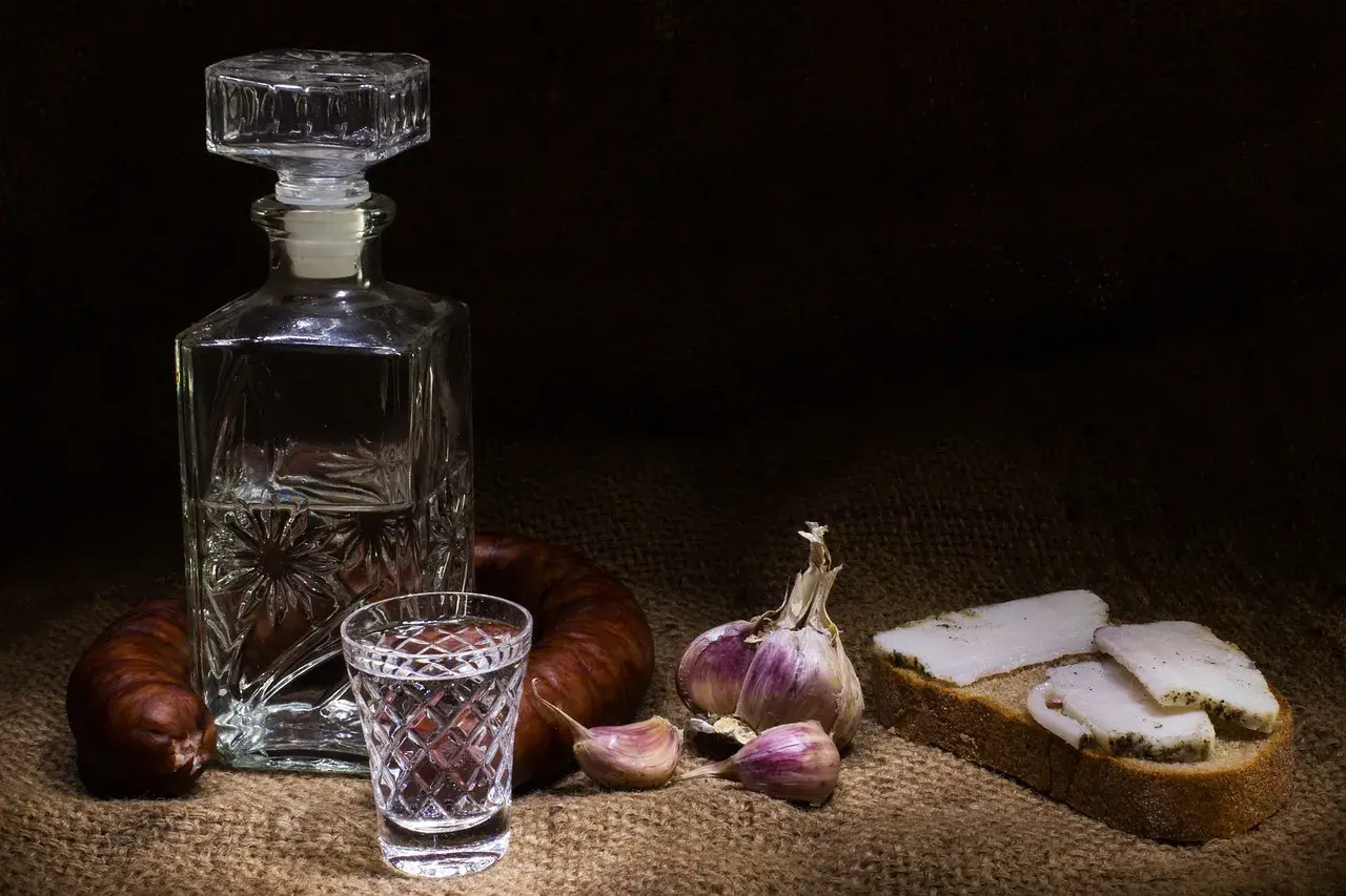 Wódka smakowa w karafce i kieliszku, obok kiełbasa, czosnek i chleb ze smalcem. Widoczne objawy utlenienia wódki smakowej po długim staniu w słońcu.