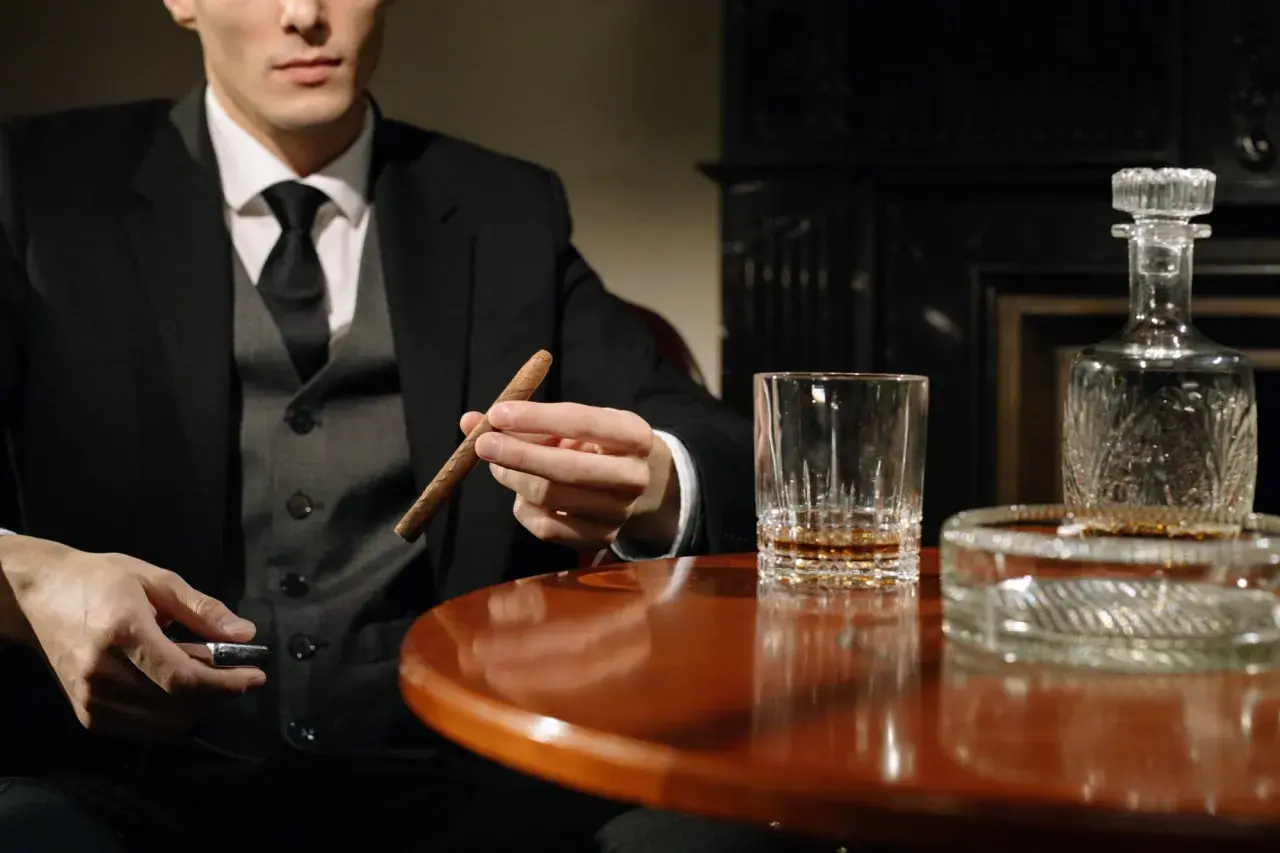 Mężczyzna w garniturze trzyma cygaro, obok szklanka whisky i karafka. Dobór whisky do cygar: intensywność i profil aromatu.