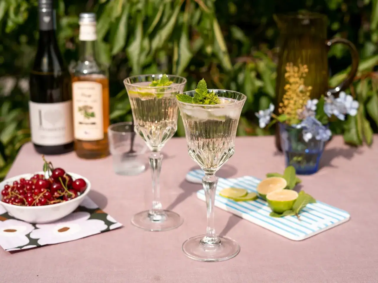 Dwa eleganckie kieliszki z orzeźwiającym drinkiem z prosecco, lodem i miętą. Obok miseczka z czerwonymi porzeczkami i cytryną.