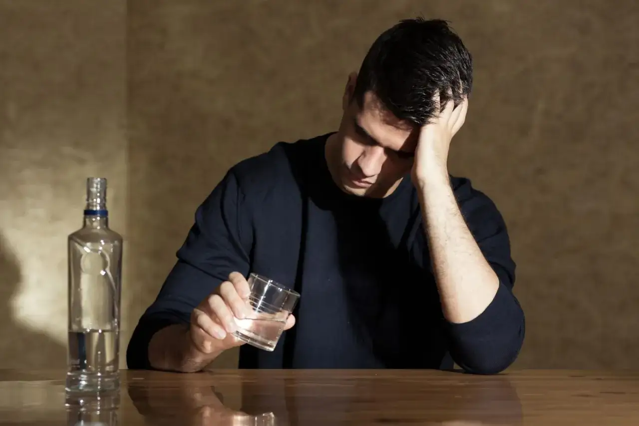 Mężczyzna z butelką alkoholu i szklanką, z ręką na czole. Objawy zatrucia alkoholem etylowym, pierwsza pomoc i środki ostrożności.