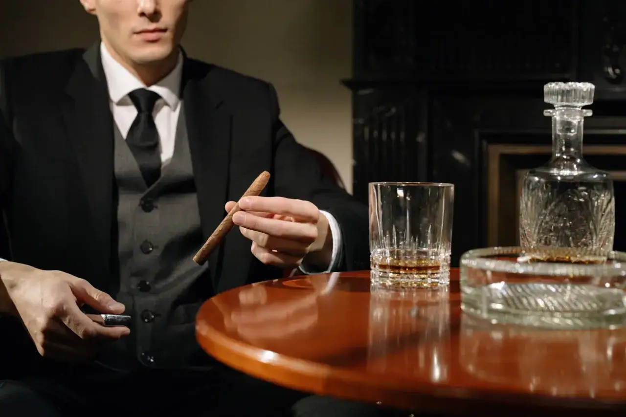 Mężczyzna w garniturze trzyma cygaro, obok szklanka whisky i karafka. Dobór whisky do cygar: intensywność i profil aromatu.