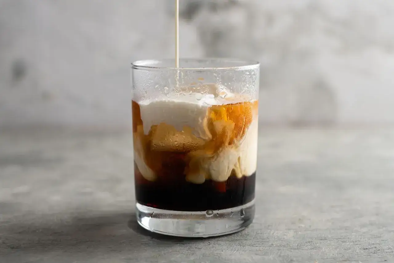 Jak dobrać wódkę do konkretnego drinka: Cosmopolitan vs White Russian. Wódka wlewana do szklanki z lodem i ciemnym płynem, tworząc efektowne smugi.
