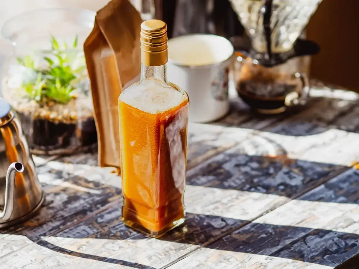 Domowy likier kawowy typu Kahlua w 15 minut. Butelka z bursztynowym płynem, kawa, terrarium i czajniczek na drewnianym blacie.