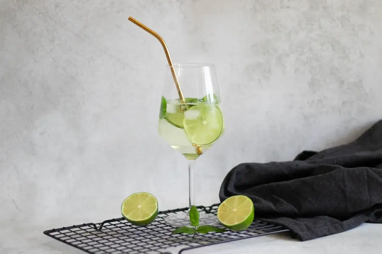 Orzeźwiający Hugo Spritz przepis: kieliszek z drinkiem, limonką, miętą i złotą słomką.