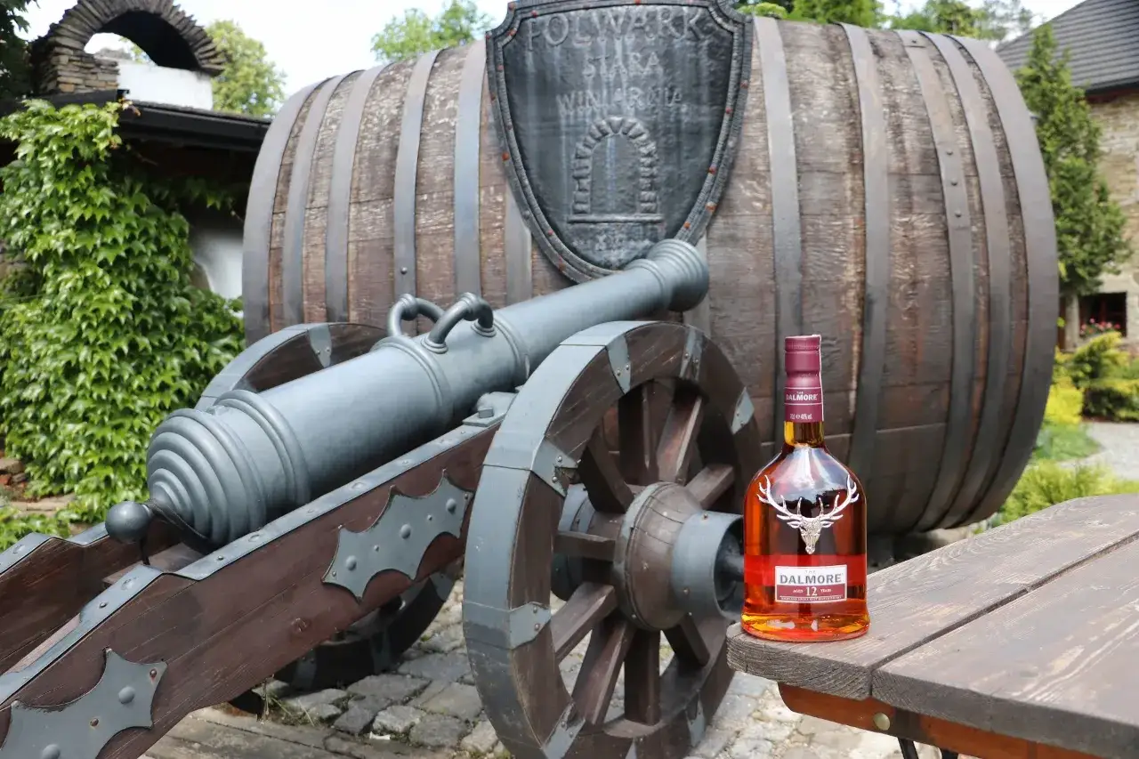 Czy warto inwestować w whisky kolekcjonerską? Portfel inwestycyjny 2026: butelka Dalmore 12 lat na tle zabytkowej beczki i armaty.