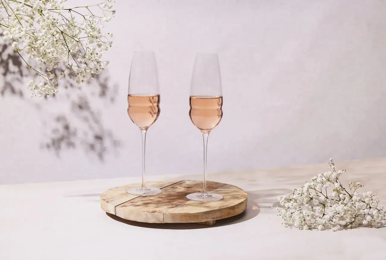 Dwa kieliszki różowego wina na drewnianej desce, otoczone drobnymi białymi kwiatami. Czy wino różowe powstaje z mieszania białego z czerwonym? Procesy produkcji są różne.