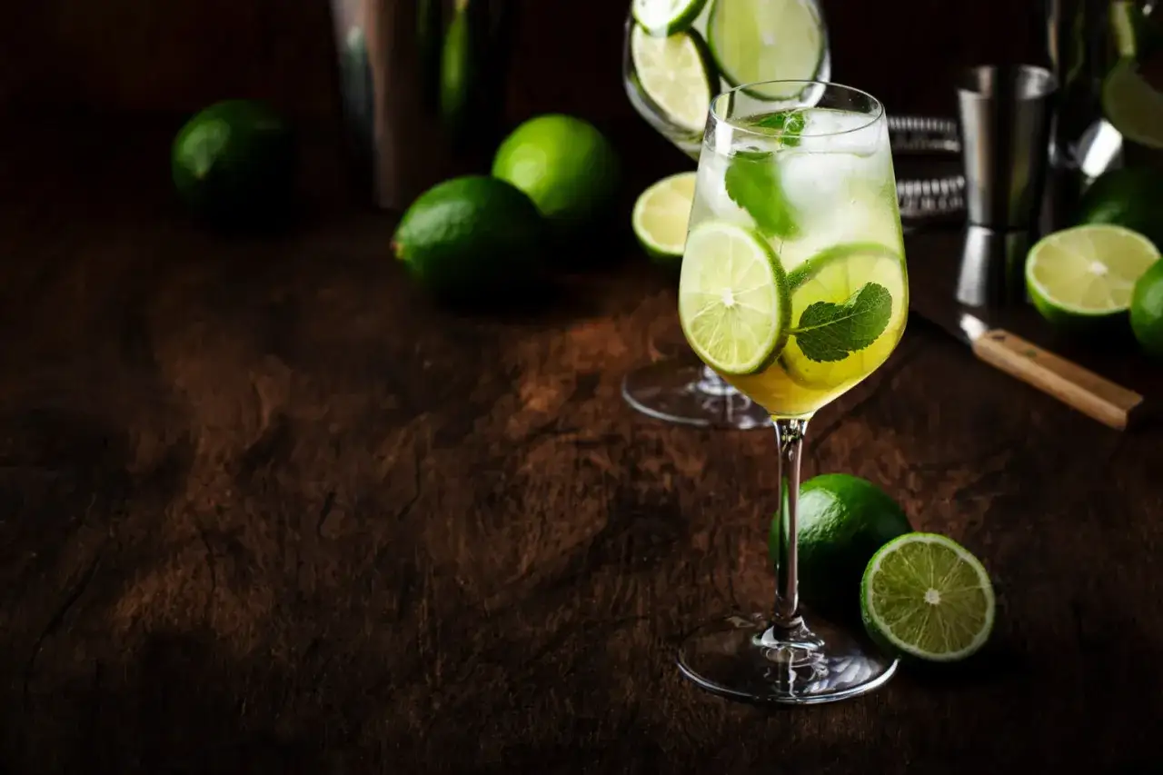 Orzeźwiający Hugo Spritz przepis. W kieliszku drink z limonką i miętą, obok świeże limonki i akcesoria barmańskie.