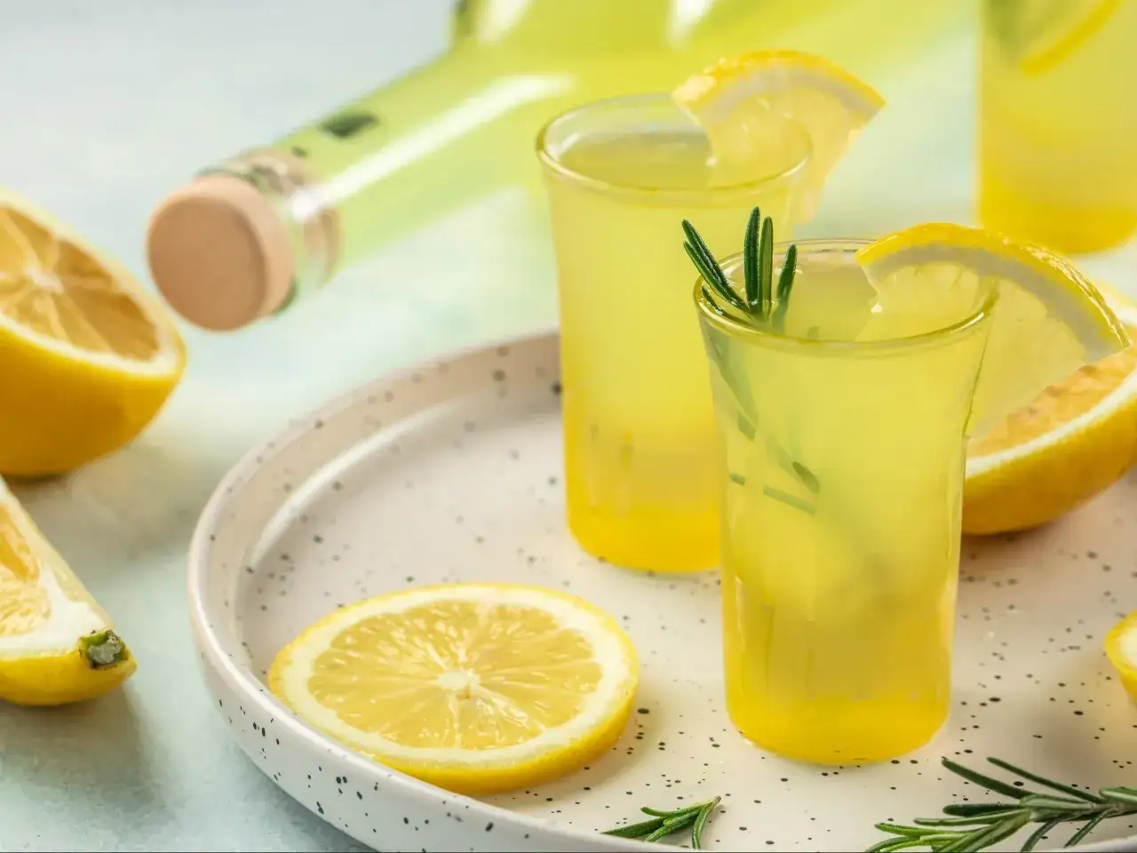 Dwa kieliszki orzeźwiającego drinka z limoncello, ozdobione plasterkami cytryny i gałązkami rozmarynu, obok butelki i świeżych cytryn.