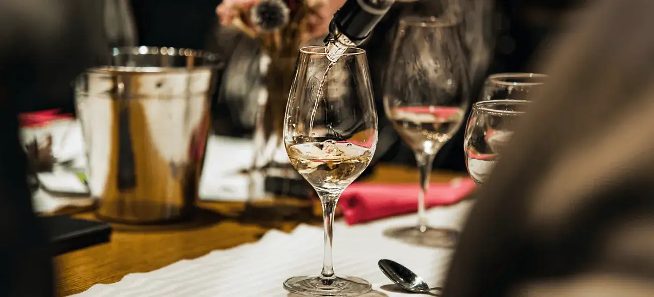 Jak zaplanować degustację wina w domu dla grupy znajomych? Nalewanie białego wina do kieliszków, obok wiaderko na lód i różowe serwetki.